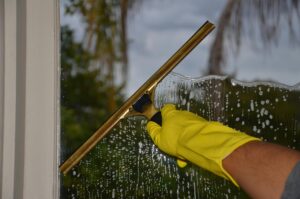 Close-up of hand cleaning window with squeegee outdoors using yellow gloves.